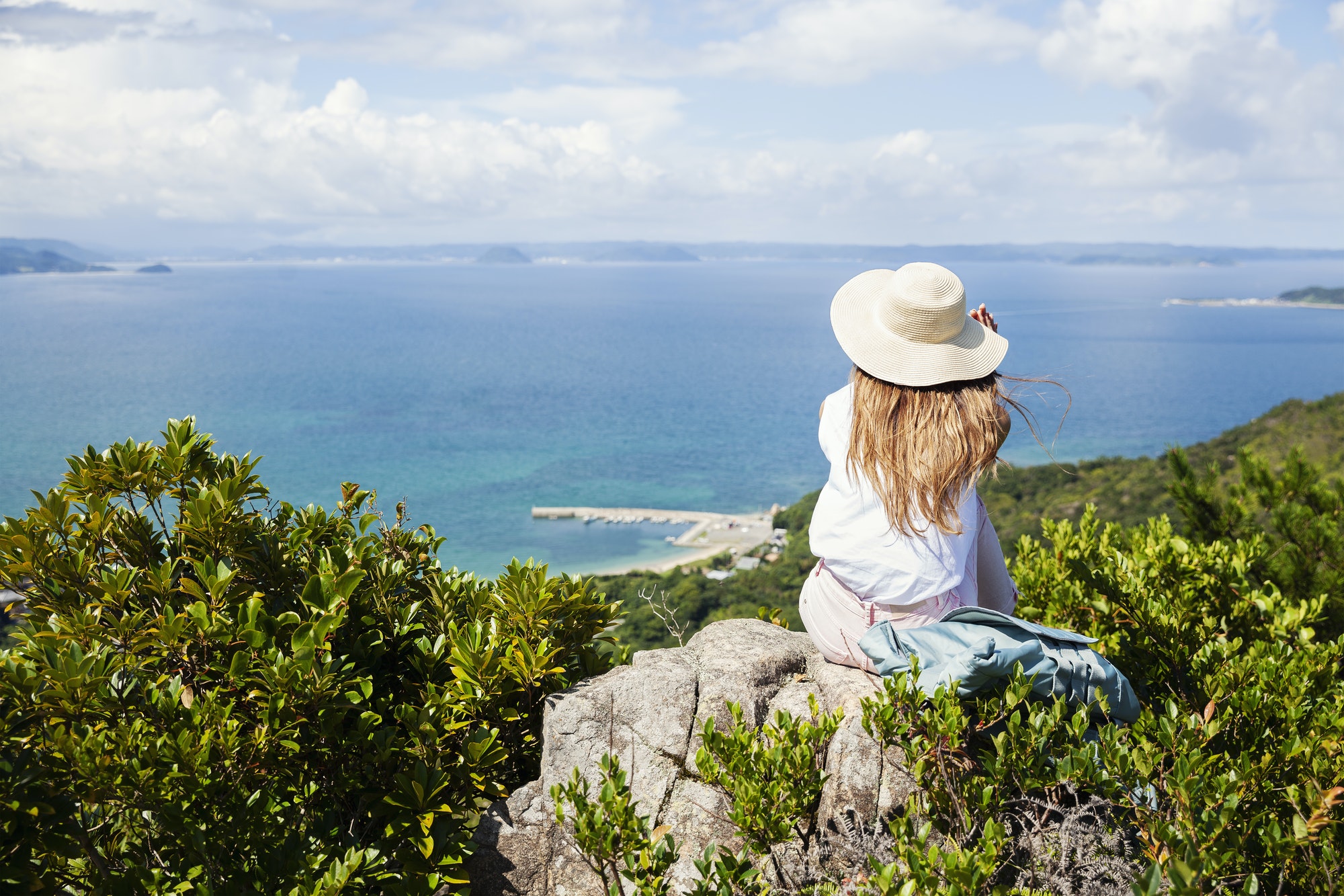 Japanese woman wearing hat sitting on rock on a cliff, ocean in the background.