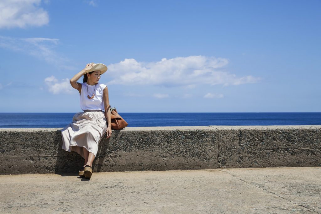 Japanese woman wearing hat sitting on a wall, ocean in the background.